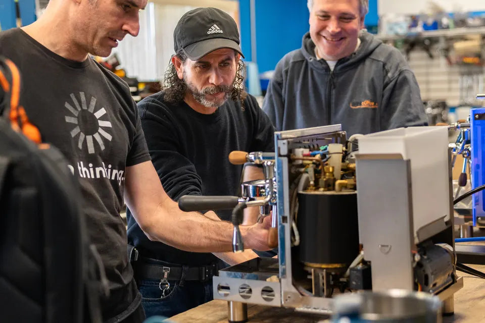 An iDrinkCoffee technician working on an espresso machine with its side panel off at the Milton service bench, two colleagues looking on